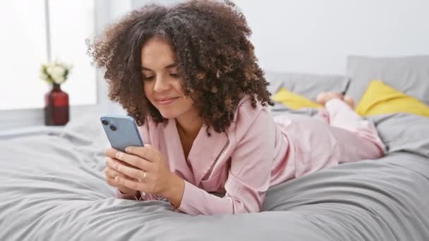 Mujer hispana alegre con el pelo rizado acostado en la cama, sonriendo felizmente mientras celebra lograr su victoria con expresión victoriosa, usando smartphone en el dormitorio - Metraje, vídeo