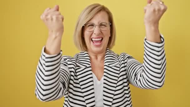 Mujer rubia extática de mediana edad con chaqueta a rayas triunfa con los brazos levantados, celebrando su alegre victoria sobre un vibrante fondo amarillo aislado. - Imágenes, Vídeo