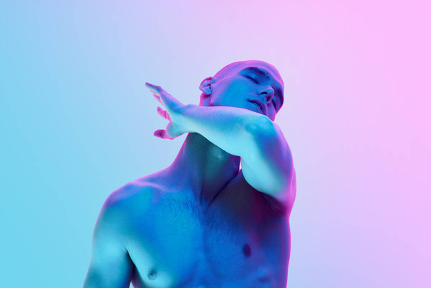 Portrait of young shirtless bald man posing with gestures on gradient blue pink background in neon light. Concept of male beauty, body, youth, fitness, sport, health - Φωτογραφία, εικόνα