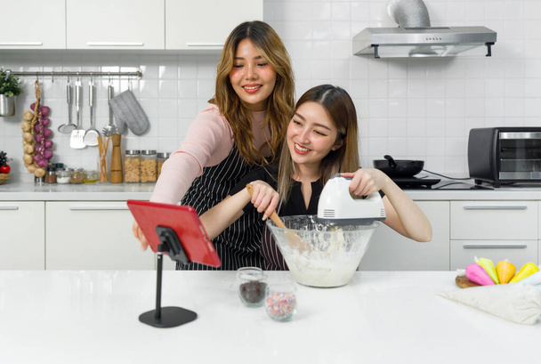 Zwei Frauen in Schürze, lächelnd und gemeinsam in einer Küche mit einem Tablet, einem Elektromixer und einer Rührschüssel backen. - Foto, Bild