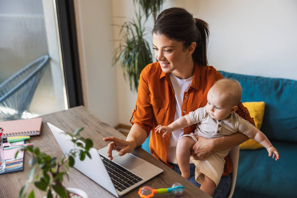 Foto de una mujer joven que trabaja en el ordenador portátil mientras cuida a su adorable bebé en casa. - Foto, imagen