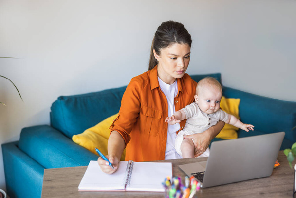 Unternehmerin schreibt Notizen, während sie am Laptop arbeitet und sich zu Hause um ihr Baby kümmert. - Foto, Bild