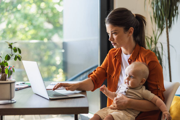 ノートパソコンで働く女性起業家と自宅で赤ちゃんの世話をする. - 写真・画像