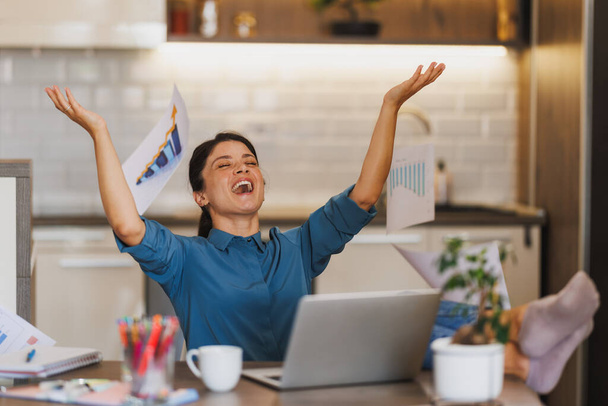 Jeune femme entrepreneure célébrant avec levé les mains dans la surprise et jetant des papiers dans les airs comme elle travaille à partir de son bureau à domicile. - Photo, image