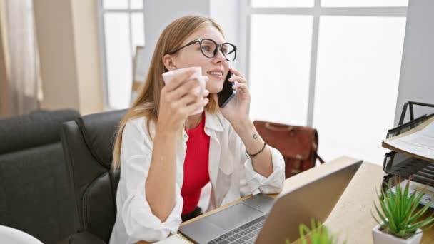 Ξανθιά γυναίκα με γυαλιά πίνει καφέ ενώ μιλάει στο τηλέφωνο στο γραφείο της.. - Πλάνα, βίντεο