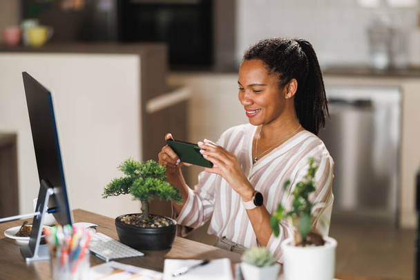 Африканская женщина владелец малого бизнеса с помощью смартфона делает фото бонсай для онлайн-продажи во время vlogging о уходе за растениями на дому. - Фото, изображение