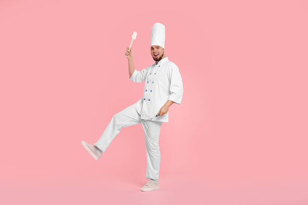 ピンクの背景にスパチュラとウィスクを保持するユニフォームで幸せなプロの菓子 - 写真・画像