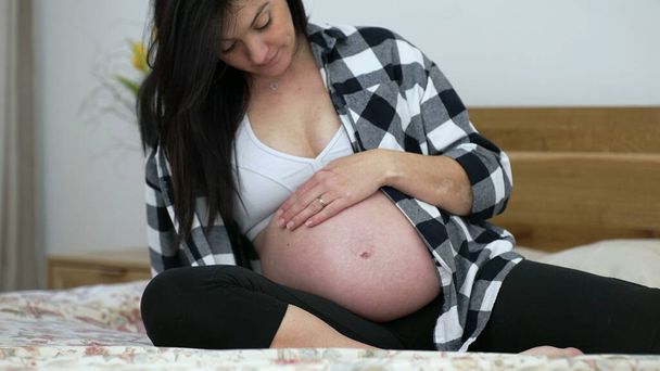 Zwangere vrouw strelen buik tijdens het derde trimester van de zwangerschap gezeten in bed, laat stadium - Foto, afbeelding