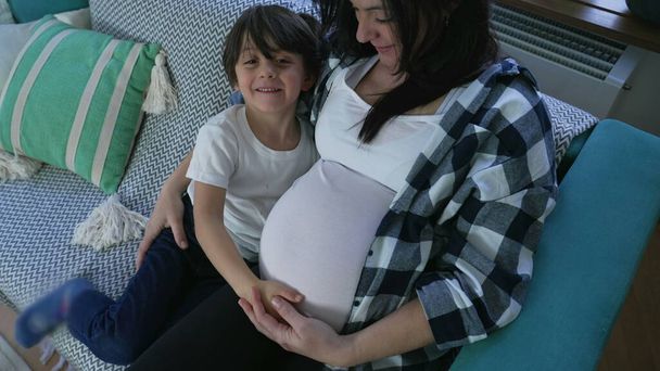 Zartes Kind umarmt schwangere Mutter aus Zuneigung zu ungeborenem Bruder, der zu Hause auf Couch sitzt - Foto, Bild