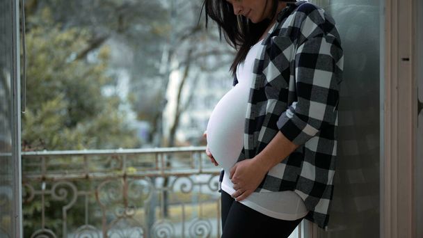 Χαρούμενη Έγκυος Γυναίκα Αγκαλιάζοντας Μητρότητα, χαϊδεύοντας με αγάπη την κοιλιά της στο Μπαλκόνι Διαμέρισμα Παράθυρο με Inspiring View - Γιορτάζοντας την Πρόβλεψη της Νεογέννητης Ζωής - Φωτογραφία, εικόνα