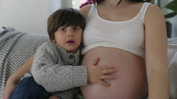 Маленький мальчик нежно трогает беременный живот матери, проявляя заботу и любовь к нерожденному младшему брату во время последней стадии беременности. - Фото, изображение