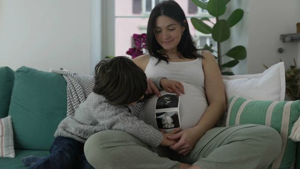 Onnellinen lapsi ja äiti istuvat kotona sohvalla hymyillen kameralle, pieni 5-vuotias poika koskettaa varovasti äidin vatsaa raskauden loppuvaiheessa, sydäntä lämmittävä todellinen perhehetki. - Valokuva, kuva