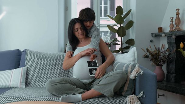 Äiti ja lapsi poseeraa kameralle, jossa näkyy syntymättömän vauvan ultraääni kolmannen raskauskolmanneksen aikana, 30s äiti hänen 5-vuotiaan poikansa kanssa kotona sohvalla odottamassa pikkuveljeä. - Valokuva, kuva
