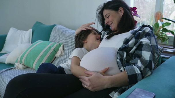 Pětiletý chlapec láskyplně objímající těhotné břicho matky na gauči, srdcervoucí rodinný moment sedící na gauči během relaxačního odpoledne. mateřský koncept - Fotografie, Obrázek