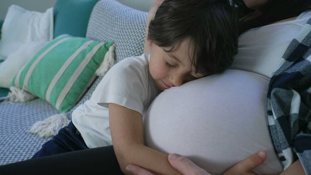 Niño de 5 años abrazando amorosamente el vientre embarazada de la madre en el sofá, momento familiar sentido sentado en el sofá durante la tarde relajante. concepto materno - Foto, imagen