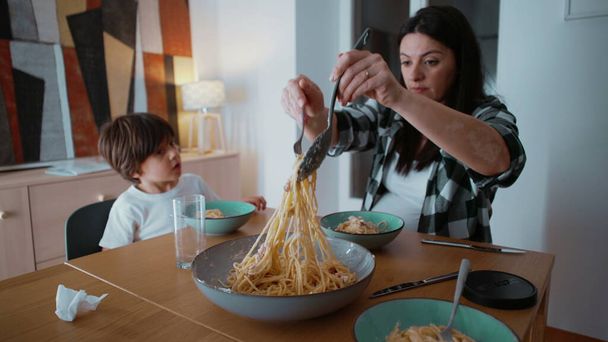 Anya és gyermeke vacsoraidőben, anya tészta spagettit szolgál fel evőeszközzel, kisfiú nézi, ahogy anya szénhidrátban gazdag ételt szolgál fel. - Fotó, kép
