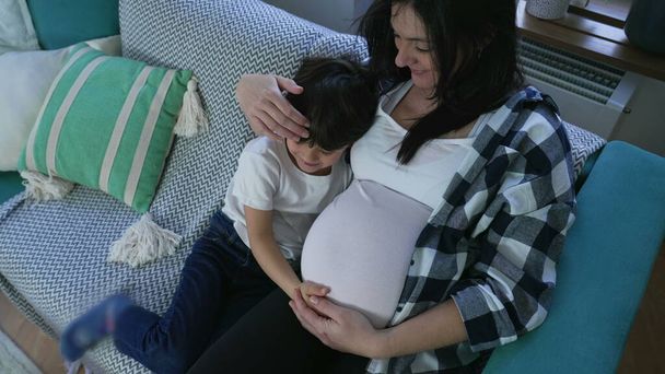 Tenero bambino che abbraccia la madre incinta in mostra di affetto al nascituro seduto sul divano di casa - Foto, immagini