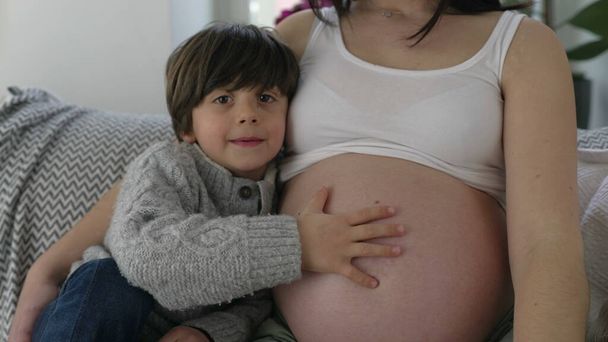 Pequeño niño tocando suavemente el vientre embarazada de la madre mostrando cuidado y amor hacia el hermano bebé nonato durante el embarazo en etapa tardía - Foto, imagen