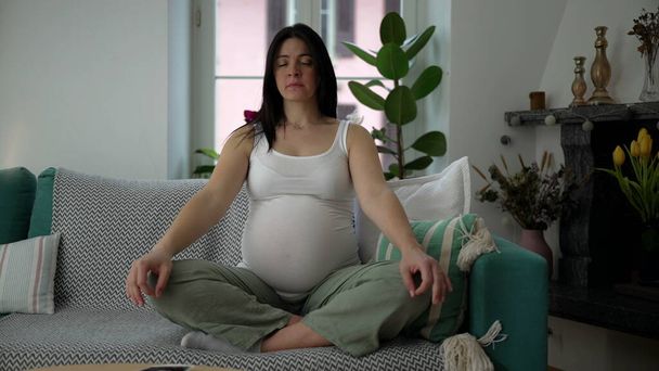 Schwangere meditiert auf der heimischen Couch mit geschlossenen Augen in besinnlicher Pose, Schwangerschaft im dritten Trimester. 30er-Jahre-Frau entspannt sich in ruhigem Appartement - Foto, Bild