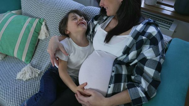 Abrazo cariñoso del niño a la madre embarazada, abrazando al hermano bebé no nacido en casa en el sofá - Foto, imagen