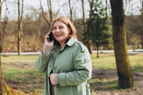 Ένα πορτραίτο μιας χαμογελαστής όμορφης γυναίκας που μιλάει στο τηλέφωνο στο πάρκο. 30s χαρούμενη γυναίκα τουρίστρια ντυμένη σε casual look ξεκουράζεται σε εξωτερικούς χώρους - Φωτογραφία, εικόνα