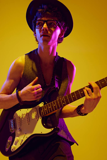 Portrait of handsome man in retro attire playing bass guitar rock-n-roll music in neon light against vibrant yellow background. Concept of music and art, hobby, concerts and festivals, modern culture - Photo, Image