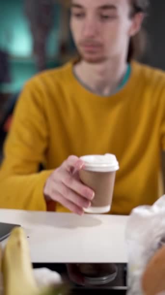 Young long haired man drinking coffee sitting before laptop among leftovers, rubbish and clothes on background. Vertical video - Footage, Video