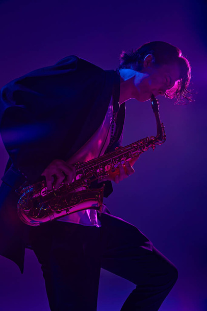 Mladý muž, talentovaný, charismatický sólista v černém obleku hrající saxofon v zářivém růžovém neonovém světle na pozadí tmavého studia. Koncept hudby, umění, hobby, koncerty a festivaly, moderní kultura - Fotografie, Obrázek