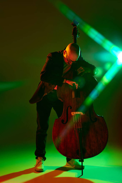 Portret łysego muzyka w okularach występującego na kontrabasie w czerwono-zielonym świetle neonowym na tle gradientowego studia. Koncepcja muzyki i sztuki, hobby, koncerty i festiwale, współczesna kultura. Ogłoszenie - Zdjęcie, obraz