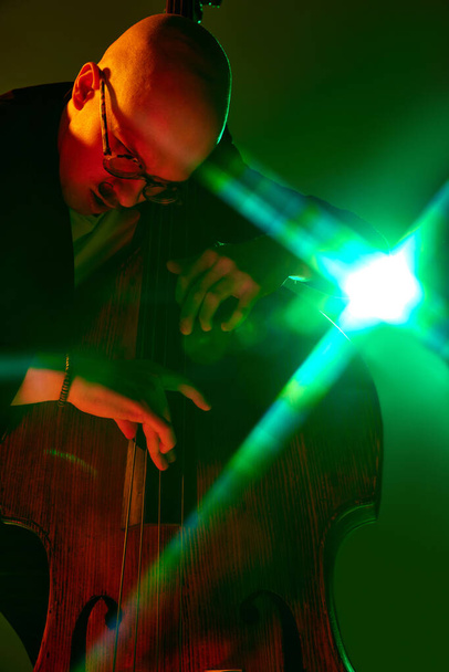 Łysy utalentowany muzyk w okularach grający na wiolonczeli w czerwono-zielonym neonie na tle gradientowego studia. Koncepcja muzyki i sztuki, hobby, koncerty i festiwale, współczesna kultura. Ogłoszenie - Zdjęcie, obraz