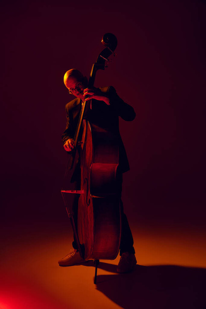 Homem careca, baixista talentoso executando composições de jazz em luz vermelho-amarela contra fundo de estúdio gradiente. Conceito de música e arte, hobby, concertos e festivais, cultura moderna. Anúncio - Foto, Imagem