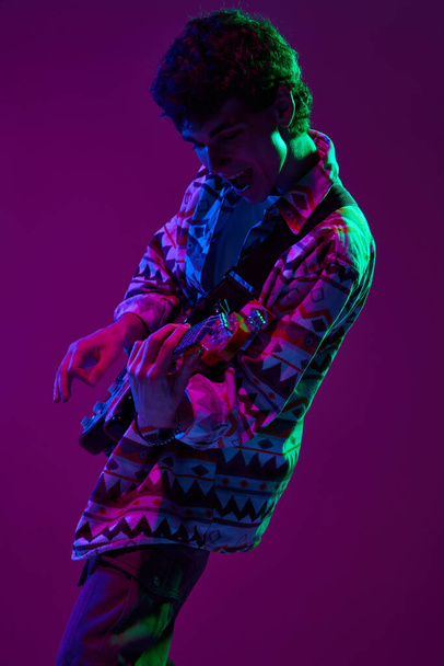 Jovem guitarrista artístico em traje retro apresentando-se em luz de néon de palco rosa-púrpura contra fundo de estúdio gradiente. Conceito de música e arte, hobby, concertos e festivais, cultura moderna. Anúncio - Foto, Imagem