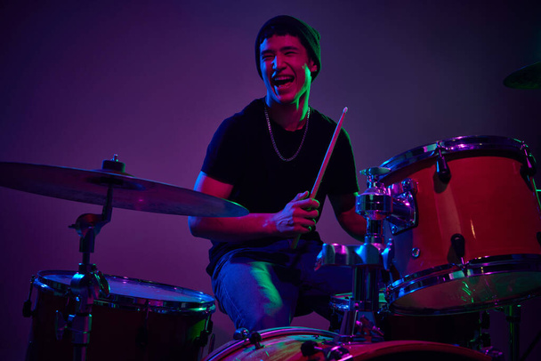 Radostný současný bubeník hraje v růžovo-fialovém jevištním osvětlení na pozadí gradientu studia. Koncept hudby a umění, hobby, koncerty a festivaly, moderní kultura. Reklama - Fotografie, Obrázek