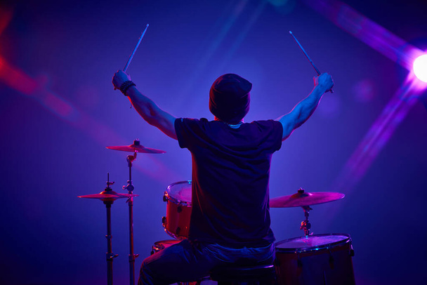 Rückansicht Porträt eines jungen Künstlers, der in rosa-lila Bühnenbeleuchtung Schlagzeug spielt, vor dem Hintergrund des Studio-Steigungsverlaufs. Konzept von Musik und Kunst, Hobby, Konzerte und Festivals, moderne Kultur. Anzeige - Foto, Bild