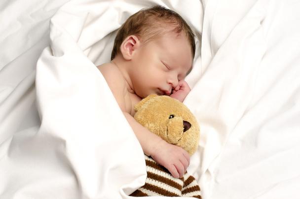 Hermoso inocente recién nacido durmiendo. Adorable niño pequeño relajándose en sábanas blancas después de un baño y sosteniendo cerca de él su juguete oso. Osito de peluche protegiendo al pequeño bebé
 - Foto, Imagen