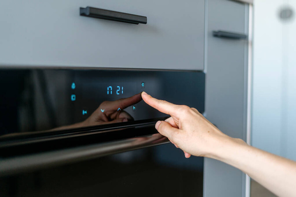 Selective focus on woman hand pressing on sensor button on domestic modern kitchen equipment for cooking food. Electric oven with control panel and timer. Blurred grey furniture background. - Photo, Image