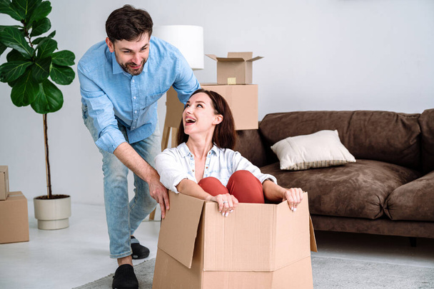 Счастливая пара веселится вместе в новой квартире в день переезда. Радостная жена сидит внутри картонной коробки и едет на полу, пока муж толкает ее в гостиной - Фото, изображение