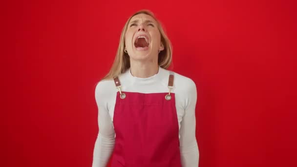 Mujer joven enfurecida, rubia y atractiva, gritando furiosamente sobre fondo rojo aislado. llevando delantal, ojos llenos de ira y frustración, rebosante agresividad - Metraje, vídeo