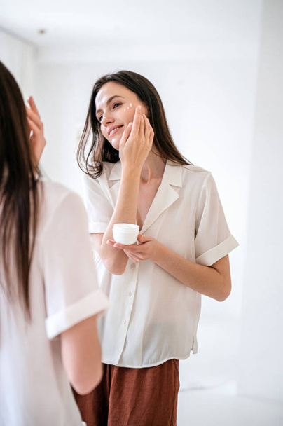 Nuori nainen sileä iho katselee peiliin, välittää hänen täydellinen kasvot ammatillinen kosmeettinen hoito. Hoitotuotteet ihonhoitotuotteet anti ikääntyminen vaikutus. Vitamiiniseerumi silmien alla - Valokuva, kuva