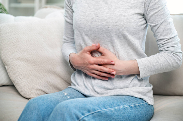 cropped shot of woman with stomach ache feeling discomfort, hold hands on belly and sitting on couch, digestive system problem. sick young female suffering from menstrual pain - Photo, Image