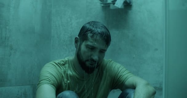 Üzüntülü sakallı yetişkin bir erkek, günlük giysiler içinde banyoda banyonun altında oturuyor. - Video, Çekim