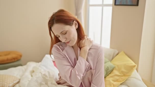 Une jeune femme caucasienne en pyjama rose s'assoit sur un lit, se frottant le cou avec une expression douloureuse dans un cadre de chambre. - Séquence, vidéo