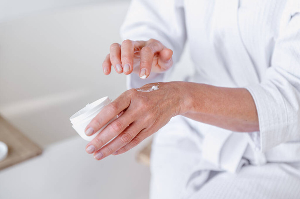 Cropped shot of mid aged woman in bathrobe applying moisturizer cream on hands for beauty skin while doing daily morning procedure. Female using anti wrinkle cosmetic product. Skincare concept - Photo, Image