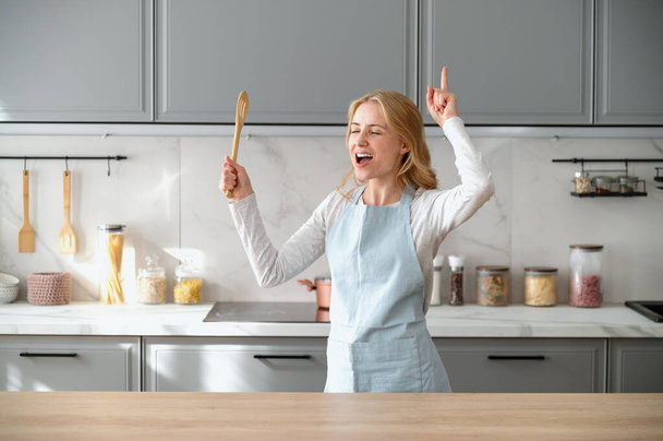 Ευτυχισμένη γυναίκα με μπλε ποδιά να χορεύει στην κουζίνα, να τραγουδάει δυνατά και να διασκεδάζει στο σπίτι. Χαρούμενη γυναίκα με ξύλινη κουτάλα στο χέρι προετοιμασία πρωινού, ακούγοντας μουσική στο διαμέρισμα - Φωτογραφία, εικόνα