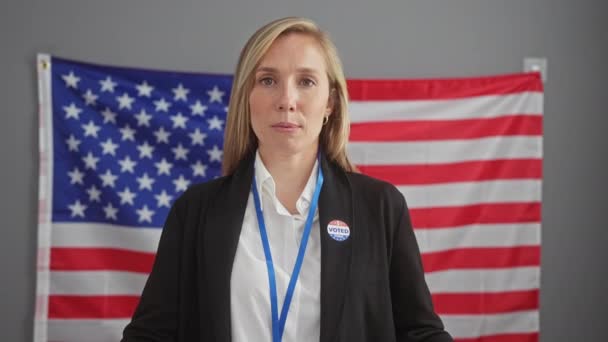 Caucasian woman in business attire with a 'voted' sticker speaks indoors against an american flag backdrop. - Footage, Video