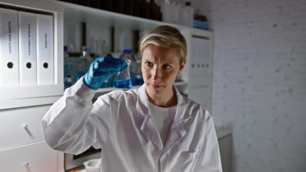 Μια στοχευμένη γυναίκα επιστήμονας εξετάζει μια μπλε χημική ουσία σε μια φιάλη σε ένα εργαστηριακό περιβάλλον. - Πλάνα, βίντεο