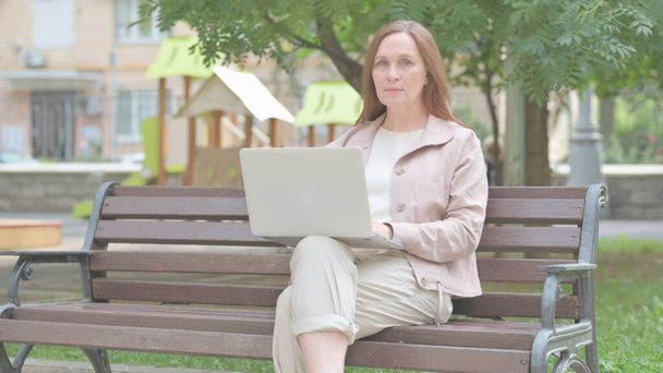 Mujer vieja moderna mirando a la cámara mientras trabaja en el ordenador portátil al aire libre - Foto, imagen