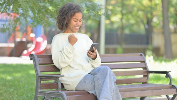 Młoda Afrykanka świętuje sukces online w telefonie podczas siedzenia na świeżym powietrzu - Zdjęcie, obraz