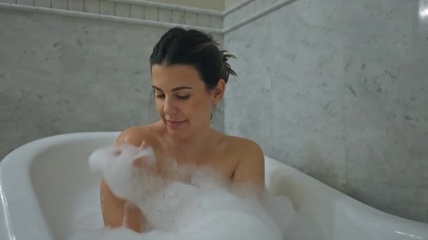 Una mujer relajada disfruta de un baño de burbujas en un moderno cuarto de baño en casa, lo que sugiere tranquilidad y cuidado personal. - Imágenes, Vídeo