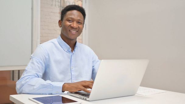 African Businessman uśmiecha się do kamery podczas pracy na laptopie w biurze - Zdjęcie, obraz